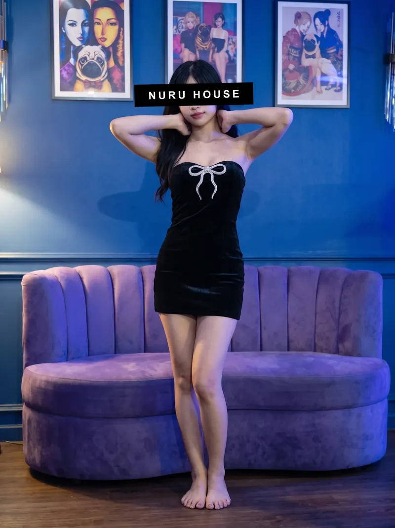 Standing in front of a round purple velvet sofa in a short black dress with a white bow, under blue light