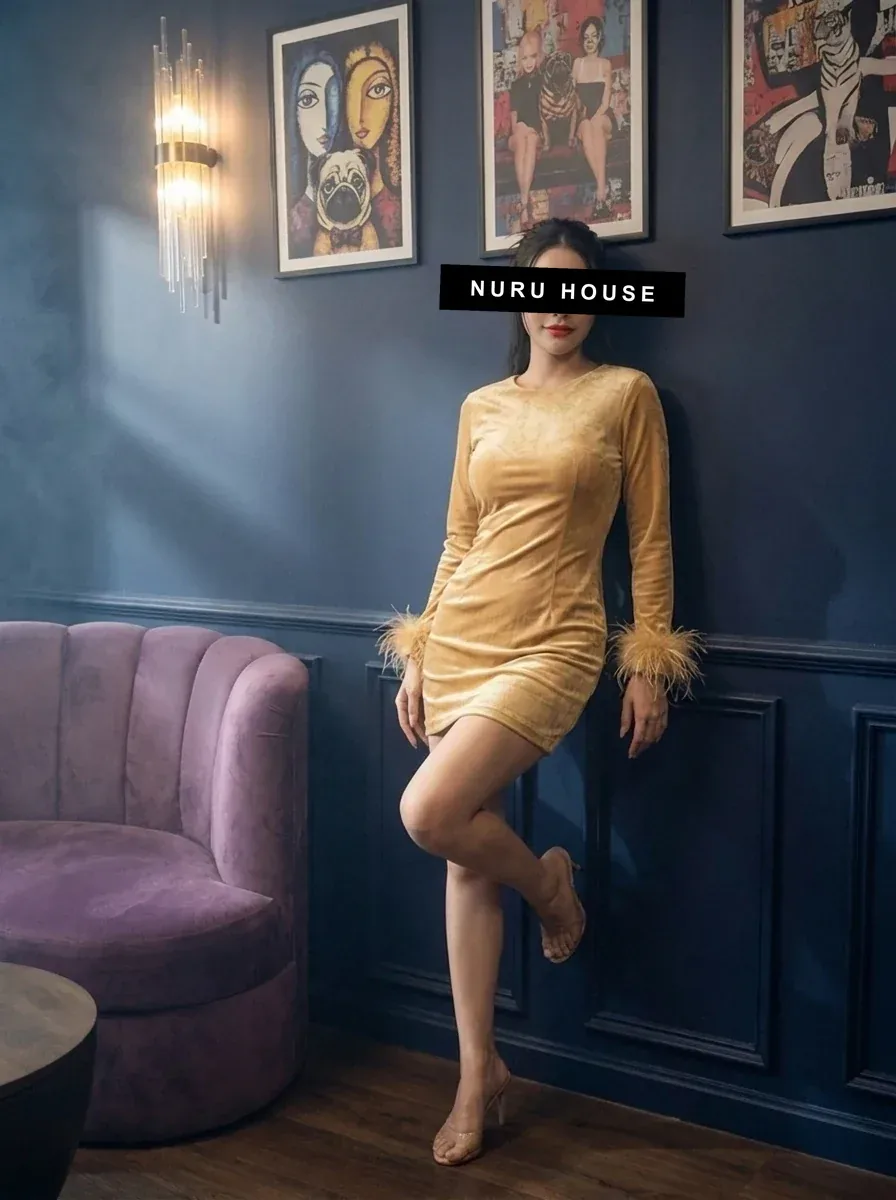 Leaning against the wall in a gold velvet mini dress with feather trim, next to a purple velvet sofa